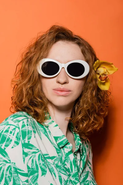 Porträt einer stilvollen rothaarigen Frau mit Orchideenblume im Haar, die mit Sonnenbrille und Bluse mit Blumenmuster auf orangefarbenem Hintergrund posiert, Sommer-Freizeit- und Modekonzept, Jugendkultur — Stockfoto