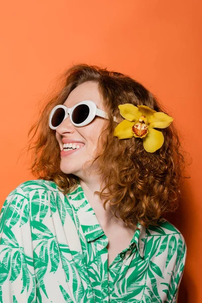 Mujer pelirroja joven feliz y confiada con flor de orquídea en el pelo, gafas de sol con estilo y blusa moderna de pie sobre fondo naranja, verano casual y concepto de moda, cultura juvenil - foto de stock