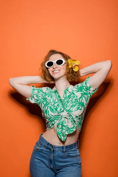 Радостная молодая рыжая женщина с цветком орхидеи в волосах и солнцезащитных очках, позирующая в блузке с цветочным узором и джинсах на оранжевом фоне, летняя повседневная и модная концепция, молодежная культура — стоковое фото
