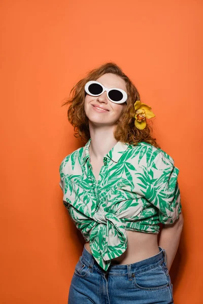 Positive junge Frau mit Orchideenblüte im roten Haar und Sonnenbrille in Jeans und Bluse mit floralem Print auf orangefarbenem Hintergrund, sommerliches Freizeit- und Modekonzept, Jugendkultur — Stockfoto