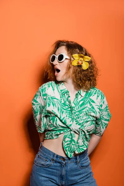 Шокированная рыжая женщина в солнцезащитных очках и цветке орхидеи в блузке с цветочным принтом и джинсах, позируя и стоя на оранжевом фоне, летняя повседневная и модная концепция, молодежная культура — стоковое фото