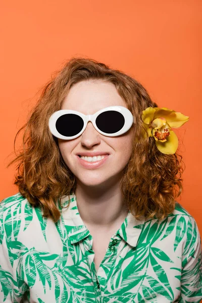 Mujer joven sonriente con flor de orquídea en pelo rojo usando gafas de sol y blusa con estampado floral mientras está de pie aislado en naranja, elegante atuendo casual y concepto de vibraciones de verano, Cultura Juvenil - foto de stock