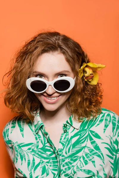 Retrato de mujer joven positiva con flor de orquídea en pelo rojo y gafas de sol mirando a la cámara mientras está de pie sobre fondo naranja, verano casual y concepto de moda, Cultura Juvenil - foto de stock