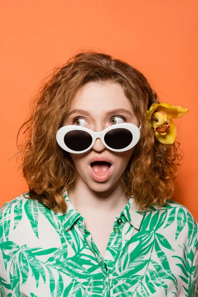 Schockierte junge Frau mit Orchideenblüte im roten Haar mit Sonnenbrille und wegschauendem Blick beim Posieren und Stehen auf orangefarbenem Hintergrund, sommerliches Freizeit- und Modekonzept, Jugendkultur — Stockfoto