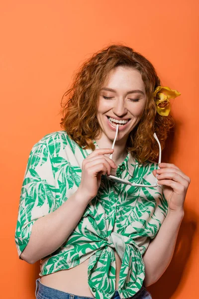Positive redhead woman with orchid flower holding fashionable sunglasses and closing eyes while standing on orange background, summer casual and fashion concept, Youth Culture — Stock Photo