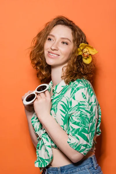 Mujer pelirroja joven sonriente con flor de orquídea en el pelo usando blusa con estampado floral y sosteniendo gafas de sol elegantes sobre fondo naranja, concepto casual y de moda de verano, cultura juvenil - foto de stock