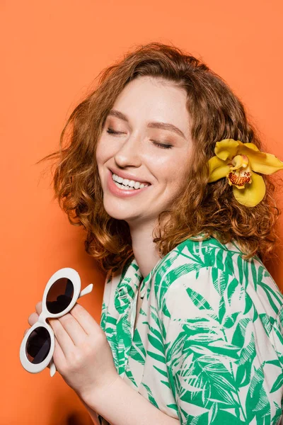 Позитивная молодая рыжая модель с цветком орхидеи в волосах, держащая солнцезащитные очки и блузку с современным цветочным узором на оранжевом фоне, летняя повседневная и модная концепция, молодежная культура — стоковое фото
