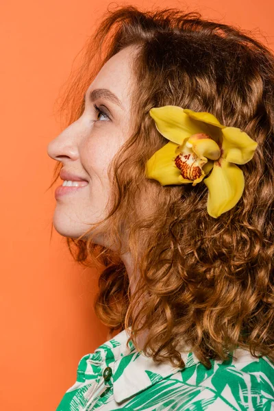 Vista lateral de la joven sonriente con flor de orquídea en pelo rojo y maquillaje natural mirando hacia otro lado mientras está de pie sobre fondo naranja, verano casual y concepto de moda, Cultura Juvenil - foto de stock