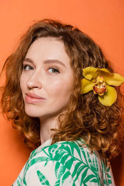 Portrait of young redhead woman with orchid flower in hair and natural makeup looking at camera while standing on orange background, summer casual and fashion concept, Youth Culture — Stock Photo