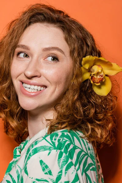 Retrato de alegre mujer joven y pelirroja con flor de orquídea en el pelo mirando hacia otro lado y posando en blusa mientras está de pie sobre fondo naranja, verano casual y concepto de moda, Cultura Juvenil - foto de stock