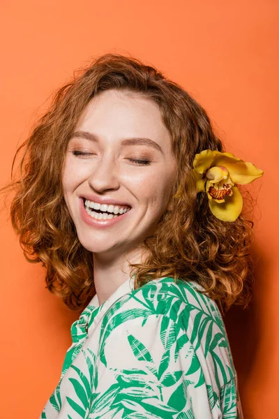 Happy young woman with orchid flower in red hair and trendy blouse with floral print standing with closed eyes on orange background, summer casual and fashion concept, Youth Culture — Stock Photo