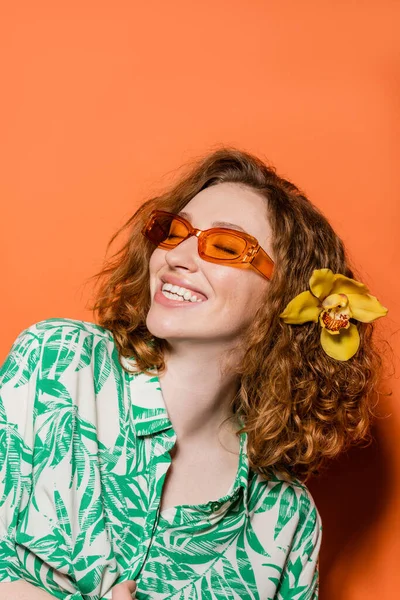 Jeune femme positive avec fleur d'orchidée en cheveux rouges portant des lunettes de soleil et chemisier élégant tout en se tenant sur fond orange, concept décontracté d'été et de la mode, Culture de la jeunesse — Photo de stock