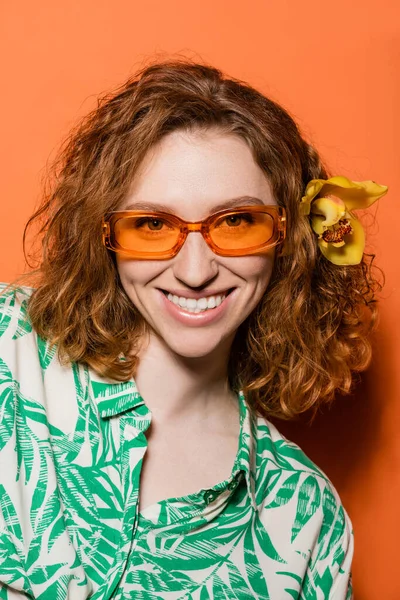 Porträt einer lächelnden und stilvollen jungen Frau mit Orchideenblüte im roten Haar, die in die Kamera blickt und mit Sonnenbrille auf orangefarbenem Hintergrund posiert, Sommer- und Modekonzept, Jugendkultur — Stockfoto