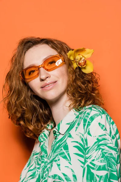 Positive young model with orchid flower in red hair, natural makeup and sunglasses posing in blouse with floral print on orange background, summer casual and fashion concept, Youth Culture — Stock Photo