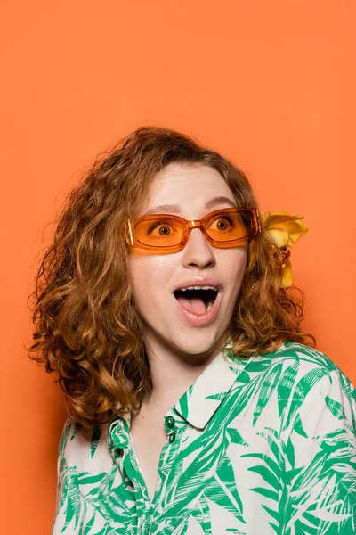 Excited young redhead woman with orchid flower in hair and sunglasses looking away while wearing blouse with floral pattern on orange background, summer casual and fashion concept, Youth Culture — Stock Photo