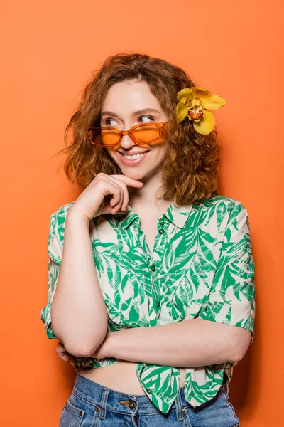 Cheerful young woman with orchid flower in hair wearing sunglasses and blouse with floral pattern while standing on orange background, summer casual and fashion concept, Youth Culture — Stock Photo