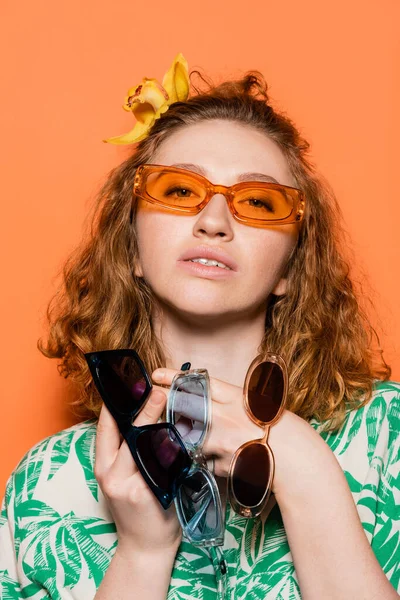 Retrato de jovem elegante com flor de orquídea em cabelos vermelhos segurando óculos de sol e olhando para a câmera enquanto estava em pé sobre fundo laranja, verão casual e conceito de moda, Cultura Jovem — Fotografia de Stock