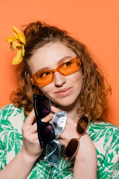 Porträt einer fröhlichen jungen Frau mit Orchideenblume im Haar, Sonnenbrille haltend und wegschauend, während sie posiert und auf orangefarbenem Hintergrund steht, Sommerleger- und Modekonzept, Jugendkultur — Stockfoto