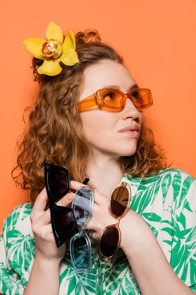 Jeune femme branchée avec fleur d'orchidée dans les cheveux tenant des lunettes de soleil et regardant loin tout en posant et debout sur fond orange, concept décontracté d'été et de la mode, Culture de la jeunesse — Photo de stock