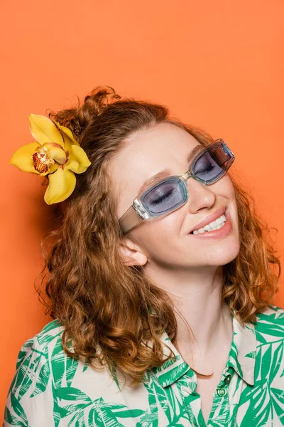 Lächelnde und trendige junge Frau mit roten Haaren und Orchideenblume posiert in Sonnenbrille und Bluse mit floralem Print auf orangefarbenem Hintergrund, Sommer-Freizeit- und Modekonzept, Jugendkultur — Stockfoto