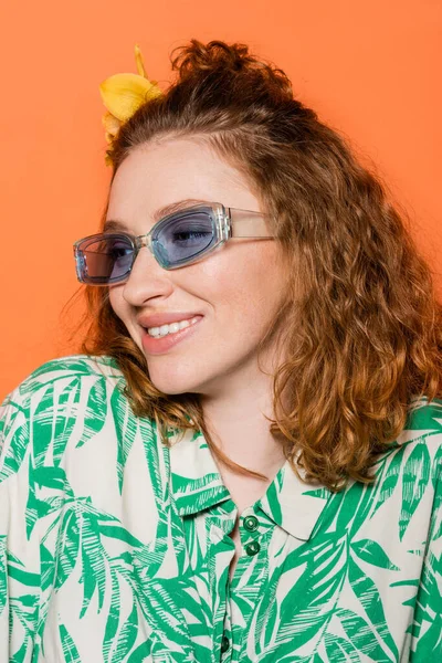 Portrait of smiling redhead woman with flower in hair and sunglasses posing in blouse with floral pattern isolated on orange, stylish casual outfit and summer vibes concept, Youth Culture — Stock Photo