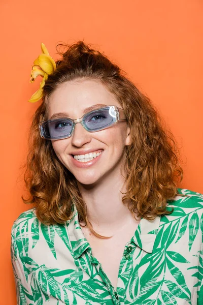 Jovem mulher ruiva alegre com flor de orquídea no cabelo e óculos de sol azuis olhando para a câmera enquanto estava isolado em laranja, roupa casual elegante e conceito de vibrações de verão — Fotografia de Stock