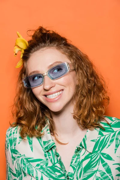 Retrato de jovem mulher ruiva alegre com flor de orquídea no cabelo e óculos de sol azuis olhando para longe e de pé sobre fundo laranja, verão casual e conceito de moda — Fotografia de Stock