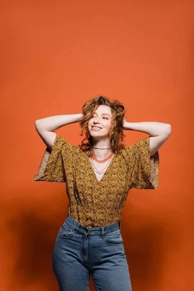 Fashionable young redhead woman in blouse with modern abstract print and jeans touching head on orange background, stylish casual outfit and summer vibes concept, Youth Culture — Stock Photo