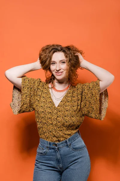 Trendy young redhead woman in modern blouse with pattern and jeans touching hair and standing on orange background, stylish casual outfit and summer vibes concept, Youth Culture — Stock Photo