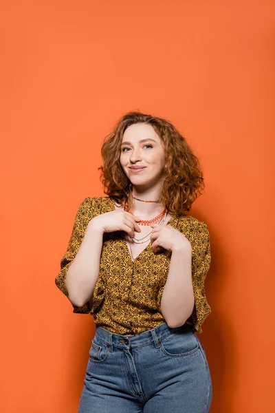 Modische und rothaarige Frau in Bluse mit abstraktem Muster und Jeans, die Halsketten berührt und auf orangefarbenem Hintergrund lächelt, stylisches lässiges Outfit und Konzept für Sommerstimmung, Jugendkultur — Stockfoto