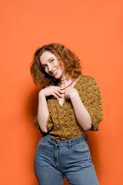 Joyful and stylish young redhead woman in blouse with abstract pattern and jeans touching necklaces and standing on orange background, stylish casual outfit and summer vibes concept, Youth Culture — Stock Photo