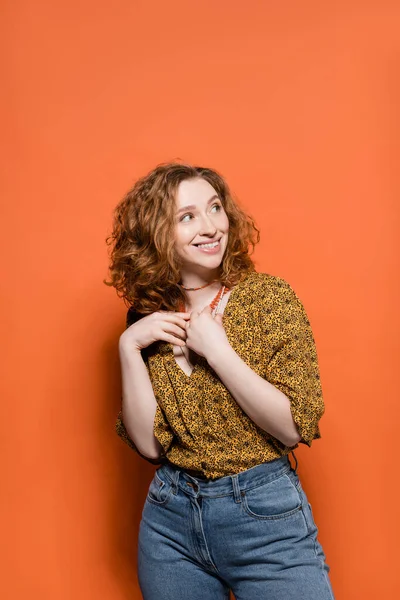 Cheerful and stylish young redhead woman in casual modern blouse and jeans touching necklaces and standing on orange background, stylish casual outfit and summer vibes concept, Youth Culture — Stock Photo