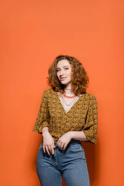 Trendy young redhead model in yellow blouse with abstract pattern and jeans smiling at camera and standing on orange background, stylish casual outfit and summer vibes concept, Youth Culture — Stock Photo