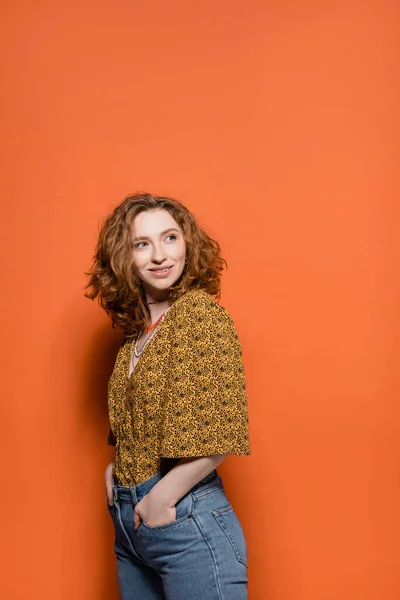 Joyful young redhead woman in blouse with abstract pattern holding hands in pockets of jeans and looking away on orange background, stylish casual outfit and summer vibes concept, Youth Culture — Stock Photo