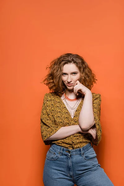 Confident and smiling red haired woman in blouse with abstract pattern and jeans looking at camera while standing on orange background, stylish casual outfit and summer vibes concept, Youth Culture — Stock Photo