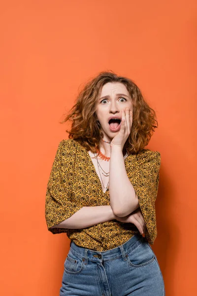 Shocked young redhead woman in blouse with abstract pattern and jeans looking at camera and standing on orange background, stylish casual outfit and summer vibes concept, Youth Culture — Stock Photo
