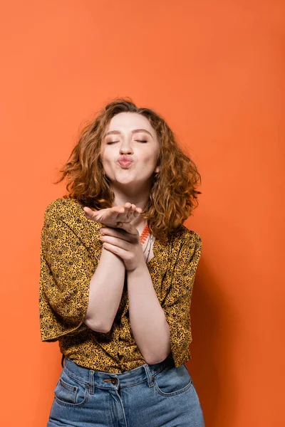 Young and stylish red haired woman in yellow blouse with abstract pattern and jeans blowing air kiss at camera on orange background, stylish casual outfit and summer vibes concept, Youth Culture — Stock Photo