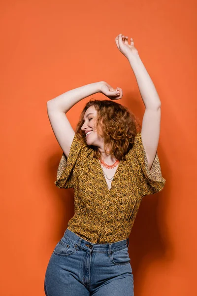 Jovem mulher ruiva alegre em blusa amarela com padrão abstrato e jeans dançando enquanto está em pé sobre fundo laranja, roupa casual elegante e conceito de vibrações de verão, Cultura Jovem — Fotografia de Stock