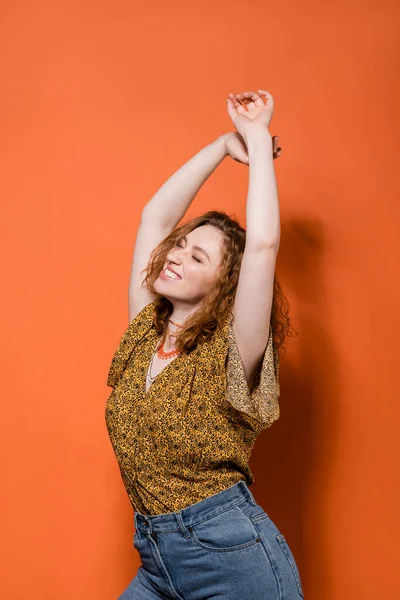 Позитивная молодая рыжая женщина в блузке с абстрактным рисунком и джинсами танцует с закрытыми глазами на оранжевом фоне, стильный повседневный наряд и летние вибрации концепции, молодежная культура — стоковое фото