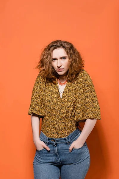 Serious young red haired model in blouse with abstract print and jeans holing hands in pockets and looking at camera on orange background, stylish casual outfit and summer vibes concept — Stock Photo