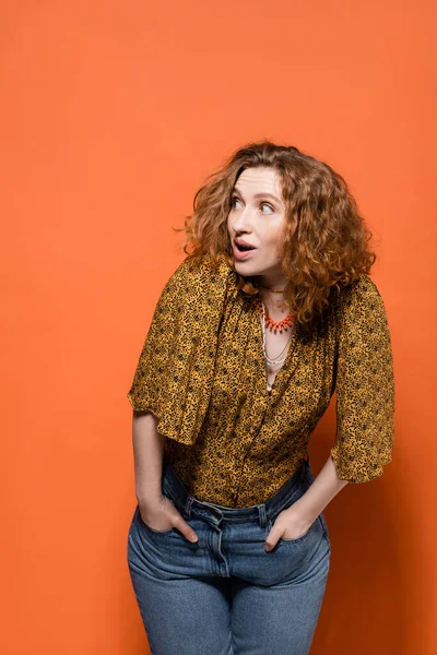Shocked young red haired woman in yellow blouse and trendy jeans looking away while posing and standing on orange background, stylish casual outfit and summer vibes concept, Youth Culture — Stock Photo