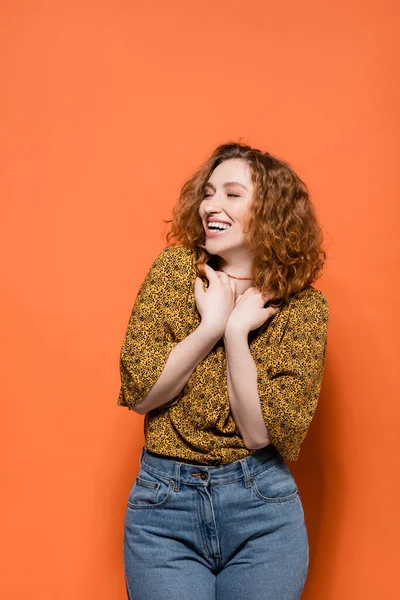 Joyful and stylish young redhead woman in yellow blouse and jeans looking away while standing on orange background, stylish casual outfit and summer vibes concept, Youth Culture — Stock Photo