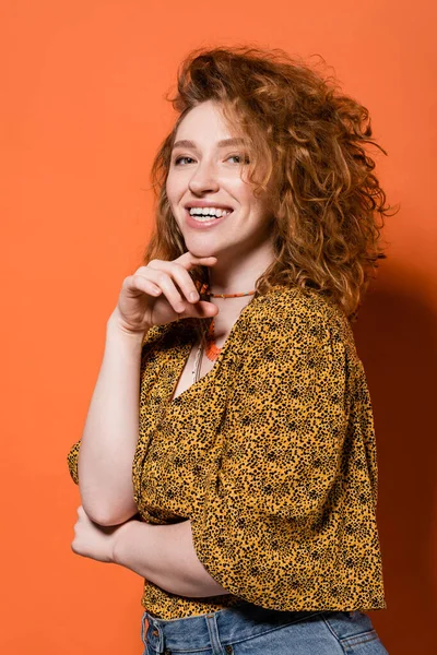 Happy young red haired woman in necklaces, yellow blouse with abstract pattern looking at camera and posing on orange background, stylish casual outfit and summer vibes concept, Youth Culture — Stock Photo