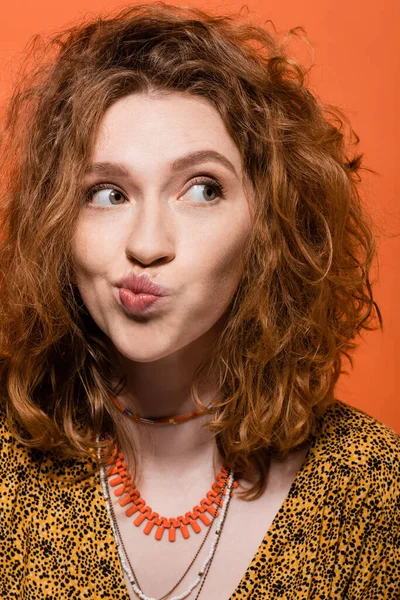 Portrait of young red haired woman in yellow blouse and necklaces pouting lips and looking away while standing isolated on orange, stylish casual outfit and summer vibes concept, Youth Culture — Stock Photo