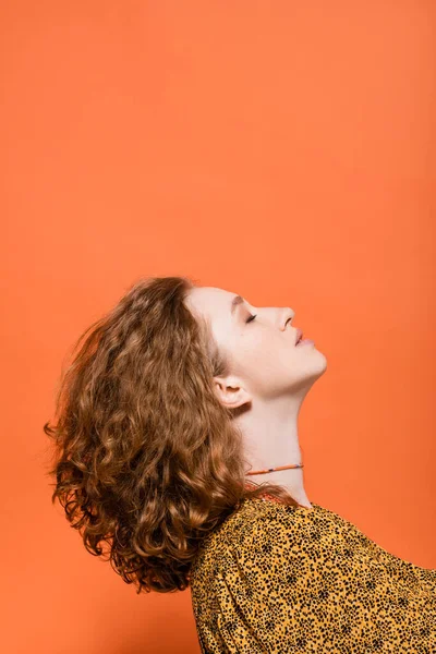 Side view of stylish young redhead woman with natural makeup in blouse with modern abstract print standing isolated on orange, stylish casual outfit and summer vibes concept, Youth Culture — Stock Photo