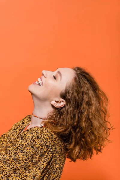 Sonriente mujer de pelo rojo joven en collares y blusa con impresión abstracta moderna divertirse y de pie aislado en naranja, elegante atuendo casual y concepto de vibraciones de verano, Cultura Juvenil - foto de stock