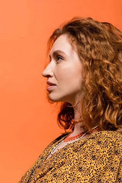 Side view of stylish redhead woman in necklaces and yellow blouse with abstract pattern looking away while standing isolated on orange, stylish casual outfit and summer vibes concept, Youth Culture — Stock Photo