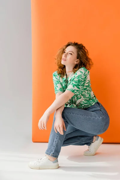 Full length of young red haired woman in blouse with floral pattern and jeans looking away while posing on orange and grey background, trendy casual summer outfit concept, Youth Culture — Stock Photo