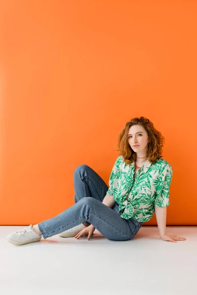Полная длина молодой рыжеволосой женщины с естественным макияжем, позируя в блузке с цветочным узором и джинсами, сидя на оранжевом фоне, модный повседневный летний наряд концепции, молодежная культура — стоковое фото