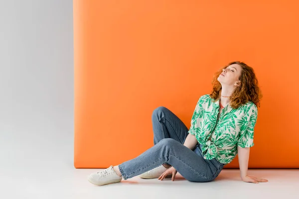 Расслабленная рыжеволосая женщина с закрытыми глазами в блузке с цветочным принтом и джинсами, сидящая на сером и оранжевом фоне, модная концепция повседневного летнего наряда, молодежная культура — стоковое фото